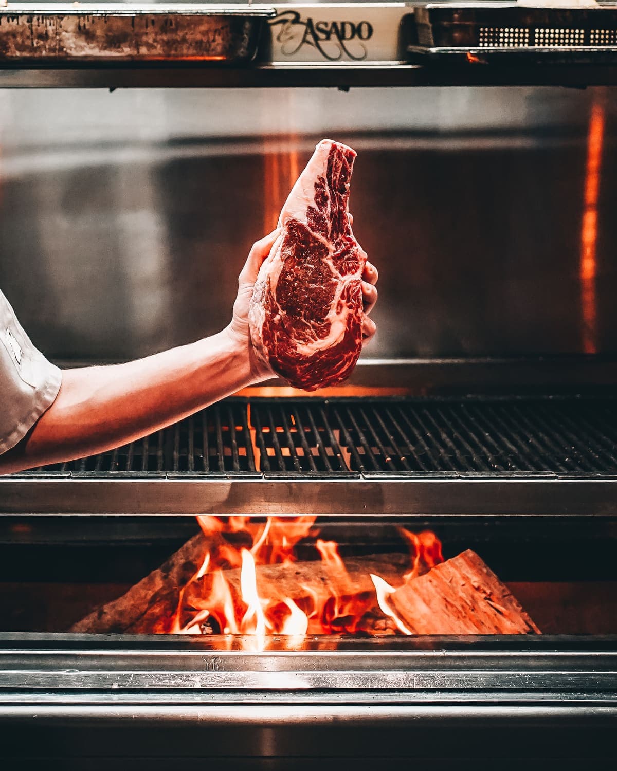 A chef holds a raw steak over the Asado grill's open flame