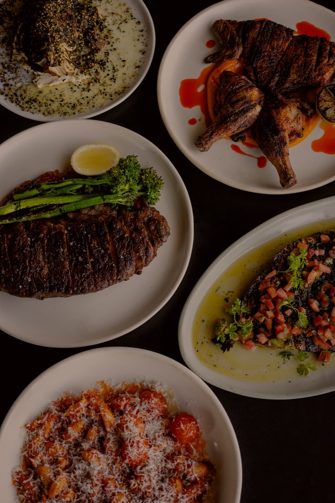 Five plates from The Inn shot from above on a dark table: whole fish, chicken off the Asado, a sliced steak with broccolini, charred eggplant, and a bowl of pasta