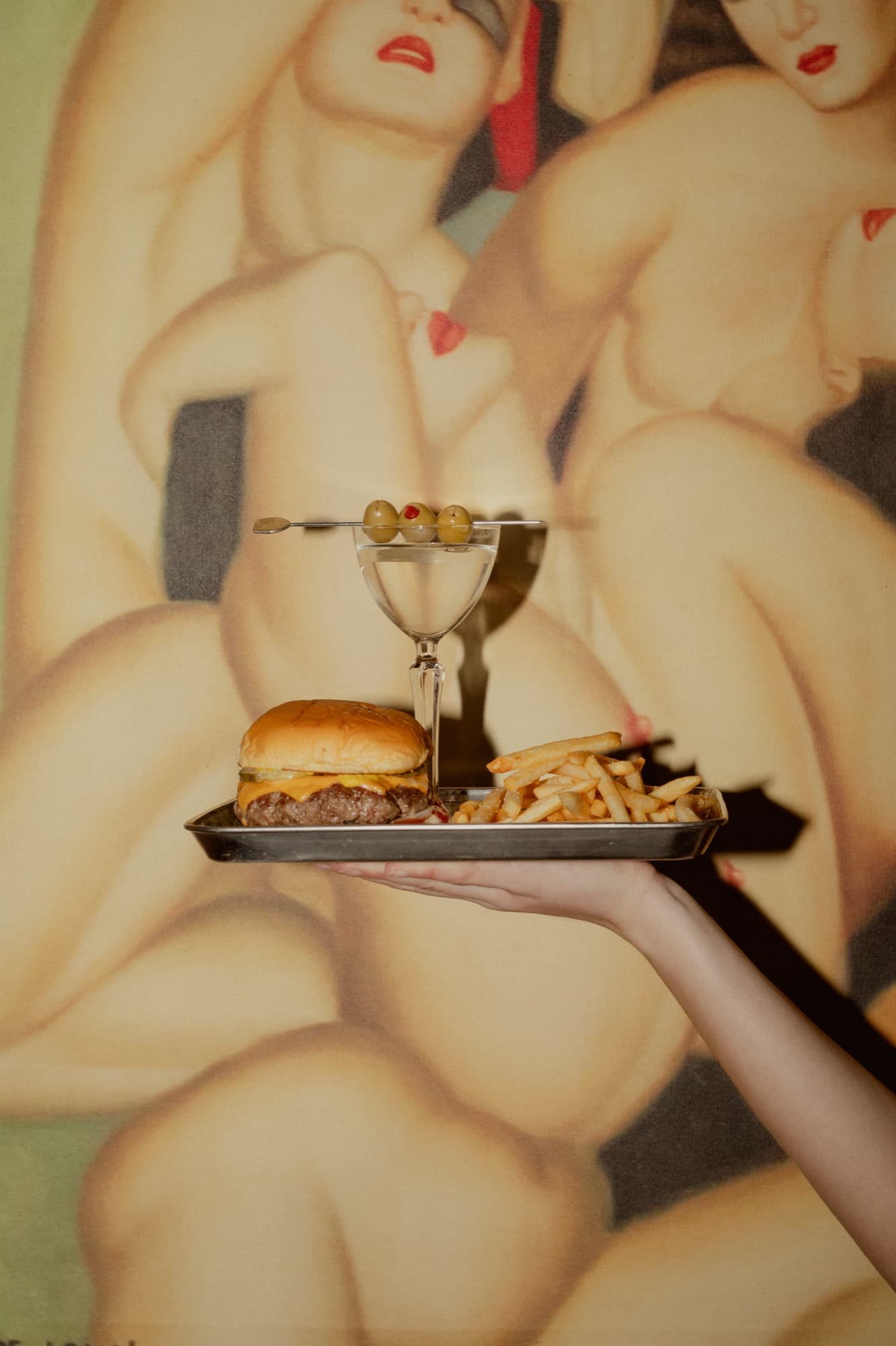 A martini with olives and a cheeseburger on a silver tray, held up against Wakefield's art-deco painting
